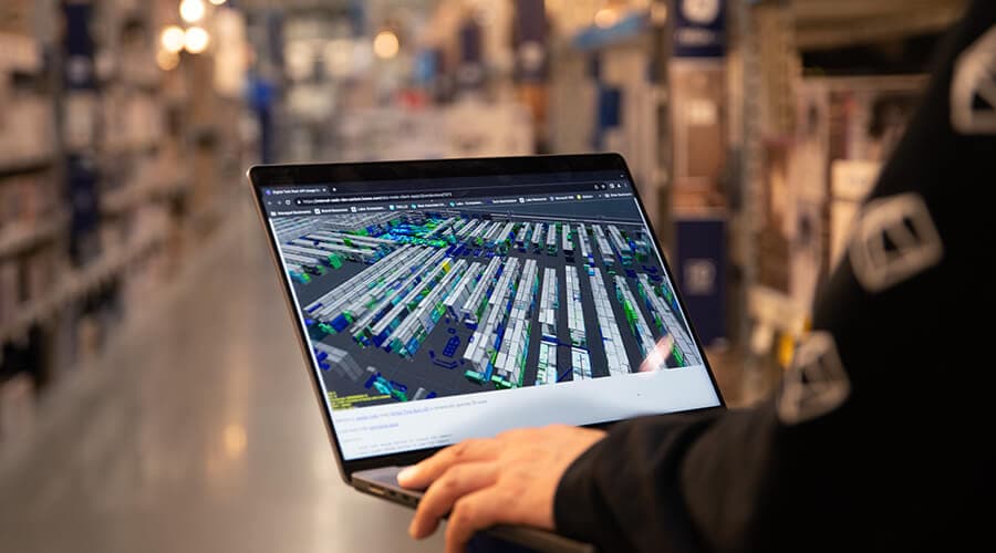 A laptop held in a warehouse aisle, showing a mapped-out view of the racking and stock around it.