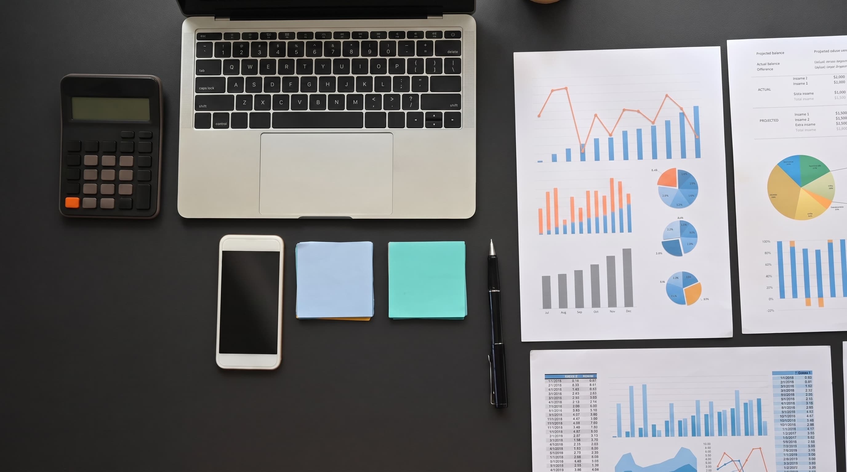 An overhead view of a laptop, calculator and phone beside printed financial charts on a desk.
