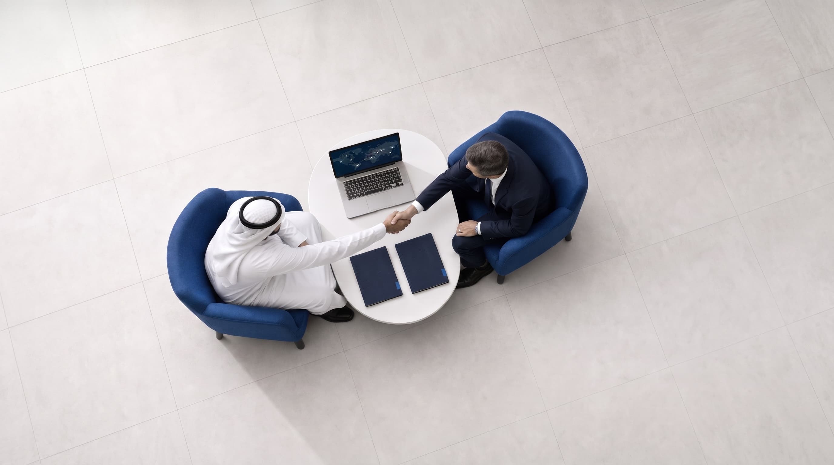 An overhead view of two businessmen shaking hands over a laptop at a round table.